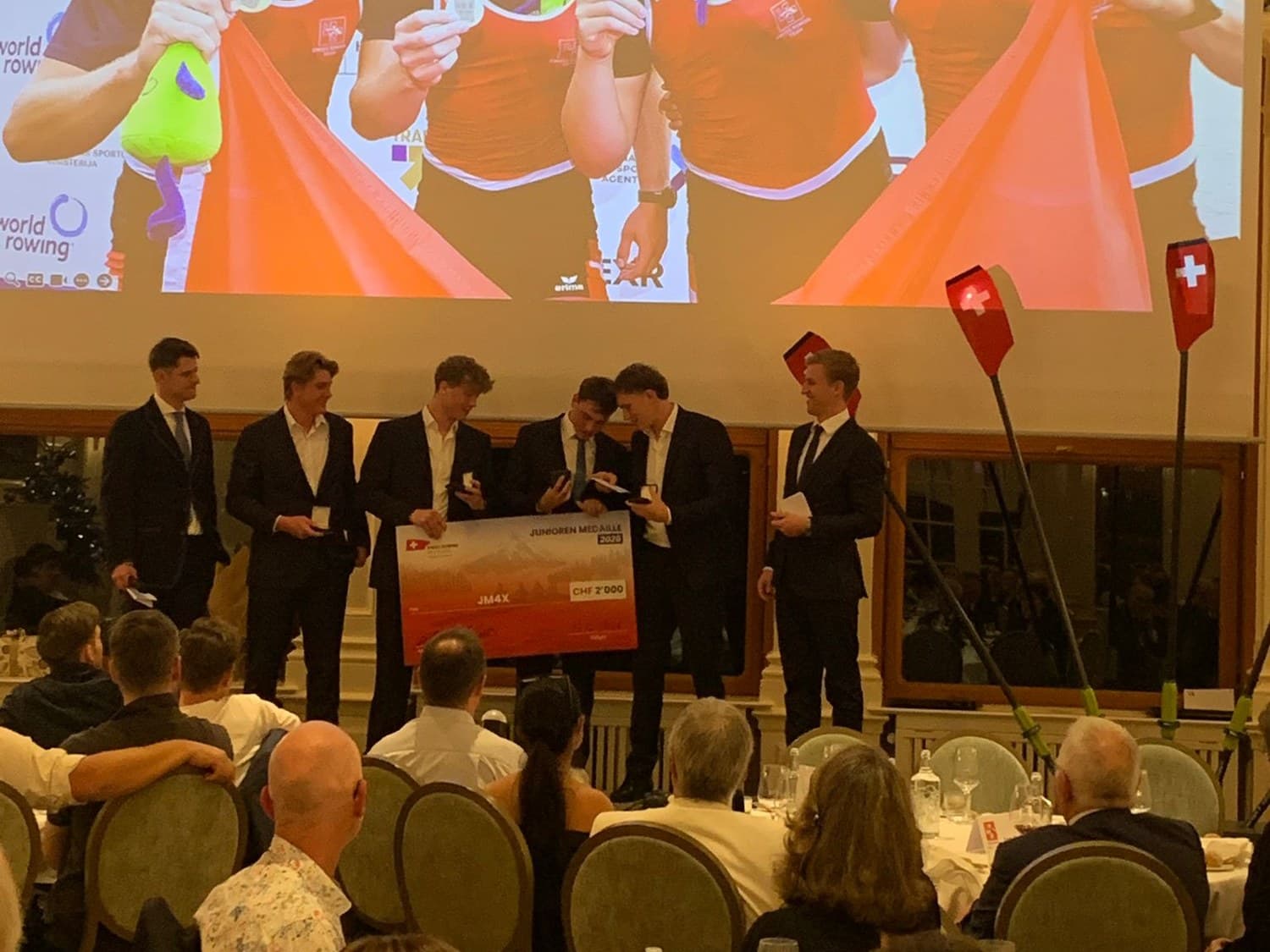 The junior crew on stage receiving the CHF 2'000 voucher at the Swiss Rowing Night, with their medal photo on the big screen behind them